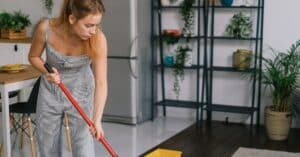 A young woman in a jumpsuit mopping a modern kitchen floor with a red mop, surrounded by plants.